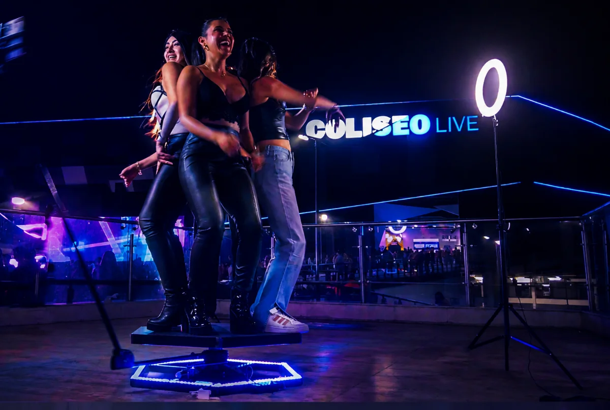 Tres amigas ríen sobre la plataforma iluminada del Video Booth 360 de Prisma en una terraza de noche